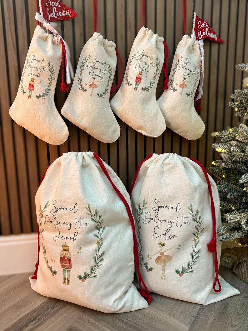May include: Five personalized Christmas gift bags with red drawstrings. The bags are made of natural canvas and feature a festive design with a wreath, stars, and a choice of a nutcracker or ballerina. The bags are labeled with the names "Jacob", "Mommy", "Daddy", "Ellie", and "Edie".