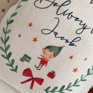 May include: A white canvas stocking with a green and red elf illustration, a red bow, and the text "Delivery to Jacob".