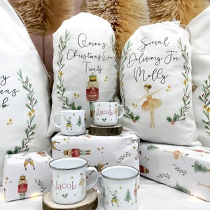 May include: Three white Christmas gift bags with personalized text, two with a nutcracker and a ballerina design, and one with a nutcracker design. The bags are sitting on a table with two white enamel mugs with a nutcracker and a ballerina design, and the name "Jacob" printed on them.