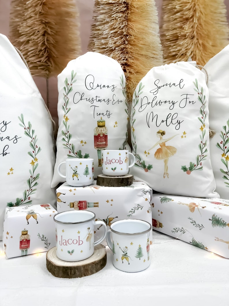May include: Three white Christmas gift bags with personalized text, two with a nutcracker and a ballerina design, and one with a nutcracker design. The bags are sitting on a table with two white enamel mugs with a nutcracker and a ballerina design, and the name "Jacob" printed on them.
