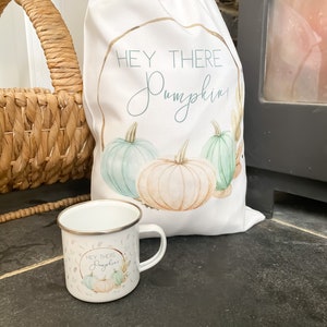 May include: A white canvas bag with the text "Hey There Pumpkin" in a gold circle and a watercolour illustration of three pumpkins. A white enamel mug with the same text and illustration is in the foreground.