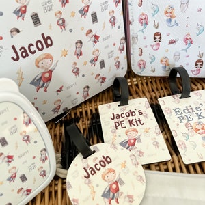 May include: A set of personalized school supplies with a superhero theme. The set includes a lunch bag, a water bottle, a PE kit bag, and a round tag. The lunch bag and water bottle feature a white background with a repeating pattern of superheroes. The PE kit bag has a white background with a superhero pattern and the text "Jacob's PE Kit". The round tag has a white background with a superhero pattern and the text "Jacob".