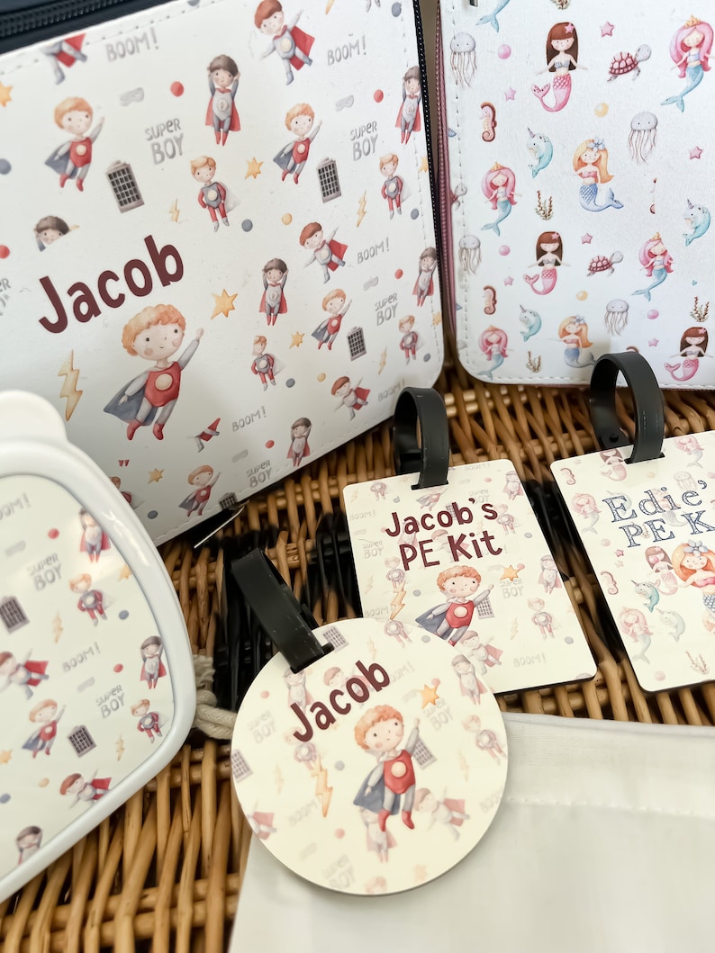 May include: A set of personalized school supplies with a superhero theme. The set includes a lunch bag, a water bottle, a PE kit bag, and a round tag. The lunch bag and water bottle feature a white background with a repeating pattern of superheroes. The PE kit bag has a white background with a superhero pattern and the text "Jacob's PE Kit". The round tag has a white background with a superhero pattern and the text "Jacob".