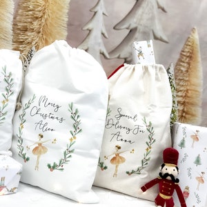 May include: Two white fabric gift bags with a ballerina and a wreath design. The bags are personalized with the names "Alice" and "Anna". One bag says "Merry Christmas" and the other says "Special Delivery For". A red toy nutcracker sits in front of the bags.