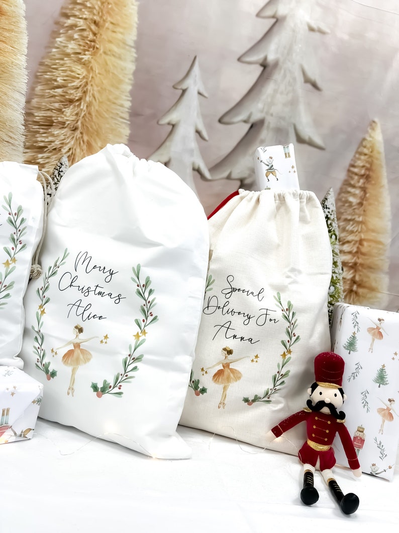 May include: Two white fabric gift bags with a ballerina and a wreath design. The bags are personalized with the names "Alice" and "Anna". One bag says "Merry Christmas" and the other says "Special Delivery For". A red toy nutcracker sits in front of the bags.