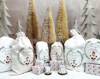 Puede incluir: Seis bolsas de regalo de tela blanca con cordones de ajuste rojos y diseños personalizados de elfos navideños. Cada bolsa tiene un nombre diferente, incluyendo "Entrega especial para Molly", "Entrega especial para Jack", "Feliz Navidad Oliver", "Entrega especial para Ruth", "Feliz Navidad Elliot" y "Entrega especial para Stephanie".