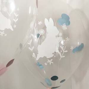 May include: Clear balloon with a white silhouette of a bunny rabbit, surrounded by white flowers and leaves. The balloon also has blue confetti inside.