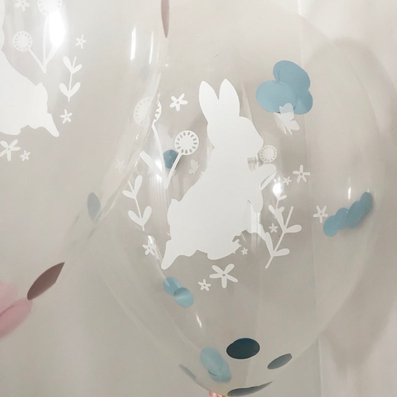 May include: Clear balloon with a white silhouette of a bunny rabbit, surrounded by white flowers and leaves. The balloon also has blue confetti inside.