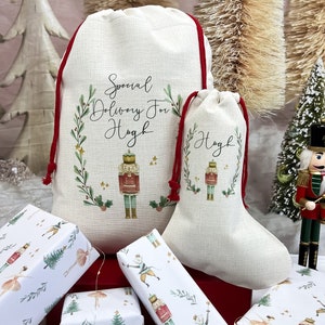 Könnte beinhalten: Zwei personalisierte Weihnachtsgeschenktüten mit roten Kordeln. Die größere Tasche trägt die Aufschrift "Special Delivery For Hugh" mit einer Nussknacker-Illustration. Die kleinere Tasche trägt die Aufschrift "Hugh" mit einer Nussknacker-Illustration. Beide Taschen sind aus natürlichem Leinenstoff gefertigt. Die Taschen sind von Geschenkpapier mit einem Nussknacker- und Ballerina-Design umgeben.