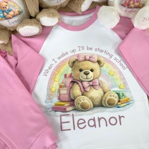 May include: A pink and white pajama set featuring a cartoon teddy bear with a backpack, bow, and the name "Eleanor." The shirt has pink sleeves and a white body with the text "When I wake up I'll be starting school." Plush teddy bears are visible.