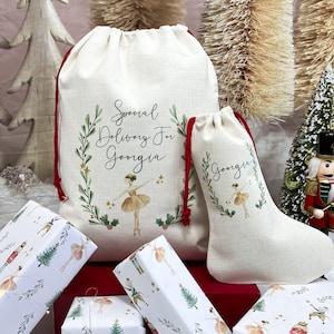 May include: Two white linen Christmas gift bags with red drawstrings. The larger bag has the text "Special Delivery For Georgia" with a ballerina illustration. The smaller bag has the text "Georgia" with a ballerina illustration. Both bags are decorated with green holly and berries.