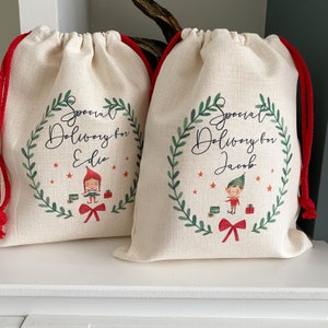 May include: Two white fabric gift bags with red drawstrings. Each bag has a green wreath with a red bow and a cartoon elf. One bag says "Special Delivery for Edie" and the other says "Special Delivery for Jacob".