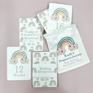 May include: A set of rainbow-themed pregnancy announcement cards with a white drawstring bag. The cards feature a rainbow, clouds, and stars. The bag has the text "Ruth's Pregnancy Journey" and "Baby Pownall Due Summer 2022".