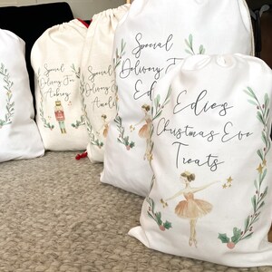 May include: Four white fabric gift bags with a Christmas theme. The bags have a green and red wreath design with a ballerina illustration. The bags are personalized with the names Aubrey, Amali, Ed, and Edie's Christmas Eve Treats.