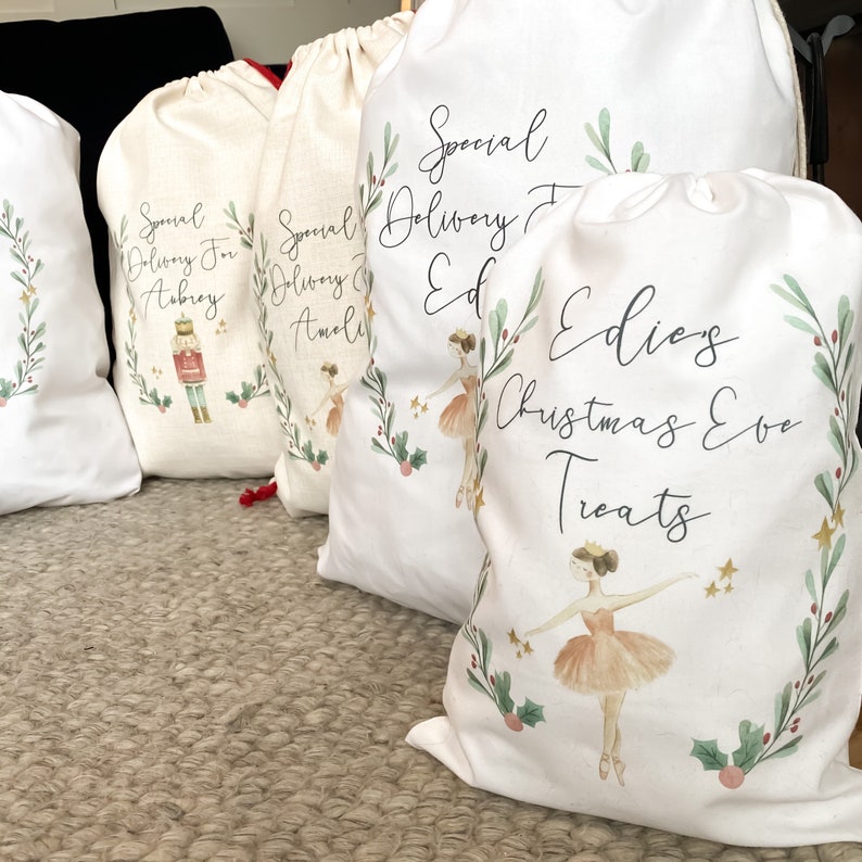 May include: Four white fabric gift bags with a Christmas theme. The bags have a green and red wreath design with a ballerina illustration. The bags are personalized with the names Aubrey, Amali, Ed, and Edie's Christmas Eve Treats.