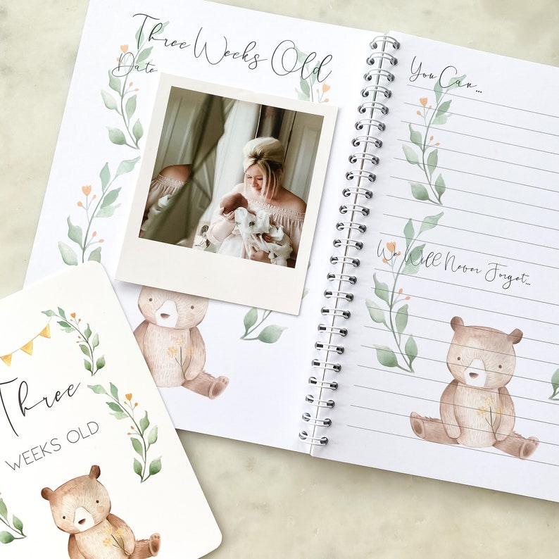 K&ouml;nnte beinhalten: Ein Baby-Erinnerungsbuch mit einer Aquarell-Illustration eines B&auml;ren und einem floralen Design. Das Buch hat eine Spiralbindung und eine Seite mit einem Foto einer Mutter, die ihr Baby h&auml;lt. Die Seite sagt "Three Weeks Old" und "You Can... We Will Never Forget..."