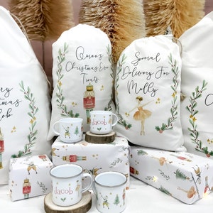 May include: Three white fabric gift bags with personalized text, two with a nutcracker and a ballerina design, and one with a Christmas Eve treats design. Two white enamel mugs with the name "Jacob" and a nutcracker design. The mugs are on wooden coasters. The bags and mugs are on a white background with a Christmas wrapping paper design.
