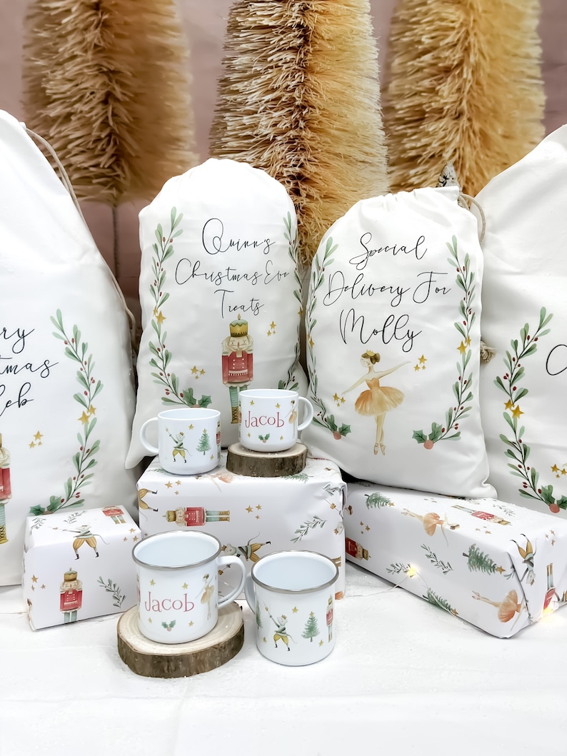 May include: Three white fabric gift bags with personalized text, two with a nutcracker and a ballerina design, and one with a Christmas Eve treats design. Two white enamel mugs with the name "Jacob" and a nutcracker design. The mugs are on wooden coasters. The bags and mugs are on a white background with a Christmas wrapping paper design.