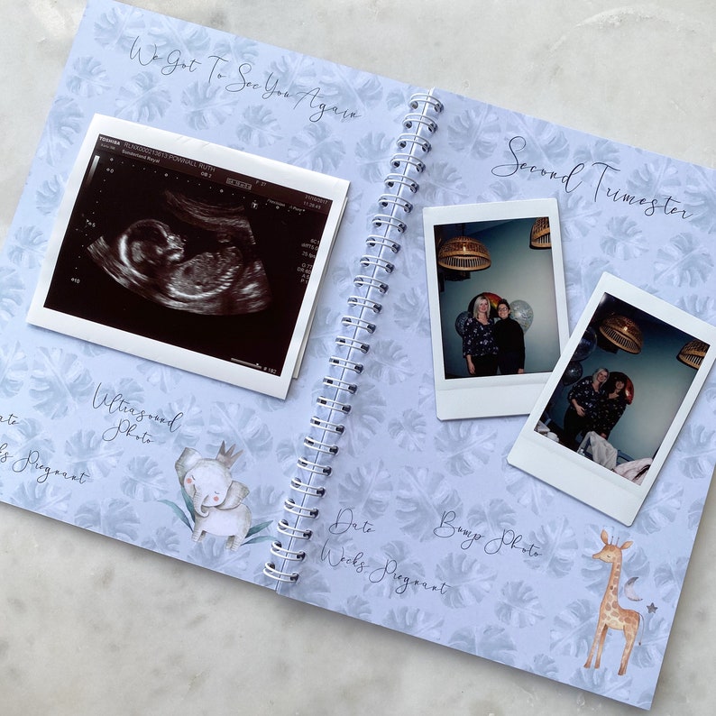 May include: A blue and white scrapbook with a floral pattern. The scrapbook has a spiral binding and includes a black and white ultrasound photo, a photo of a pregnant woman, and the text "We Got To See You Again" and "Second Trimester".