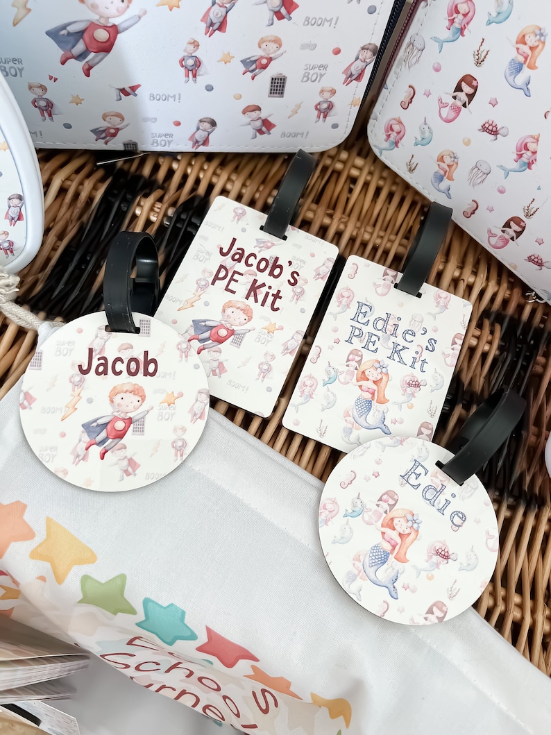 May include: Two round luggage tags with black straps. One tag features a cartoon superhero boy with the name "Jacob" and the text "Jacob's PE Kit". The other tag features a cartoon mermaid with the name "Edie" and the text "Edie's PE Kit".
