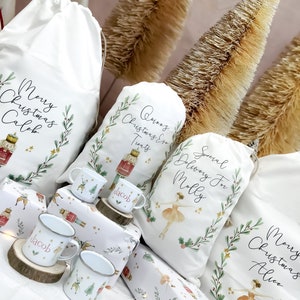 May include: A collection of white fabric Christmas gift bags with personalized names and festive designs. The bags feature illustrations of a ballerina, a nutcracker, and holly leaves. The bags are arranged on a table with wrapping paper and small mugs.