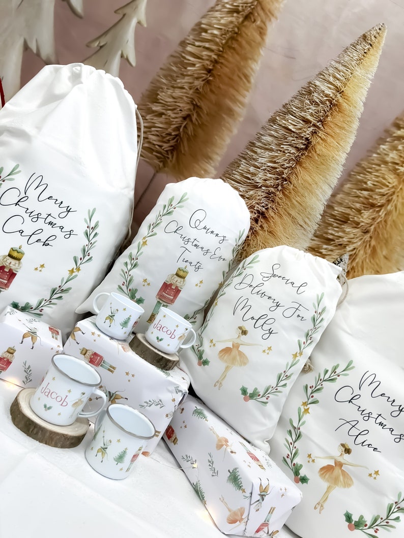 May include: A collection of white fabric Christmas gift bags with personalized names and festive designs. The bags feature illustrations of a ballerina, a nutcracker, and holly leaves. The bags are arranged on a table with wrapping paper and small mugs.