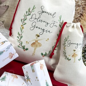 May include: Two white fabric gift bags with red drawstrings. Both bags feature a green wreath with red berries and gold stars. The larger bag has the text "Special Delivery For Georgia" and a ballerina illustration. The smaller bag has the text "Georgia" and a ballerina illustration.