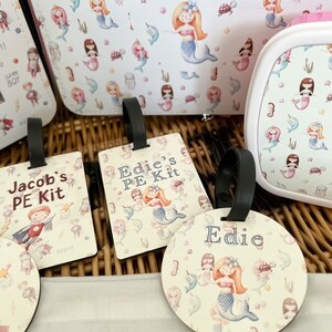 May include: Three personalized luggage tags with a mermaid theme. The tags are made of wood and have a white background with a colorful mermaid design. The text on the tags reads "Jacob's PE Kit", "Edie's PE Kit", and "Edie".