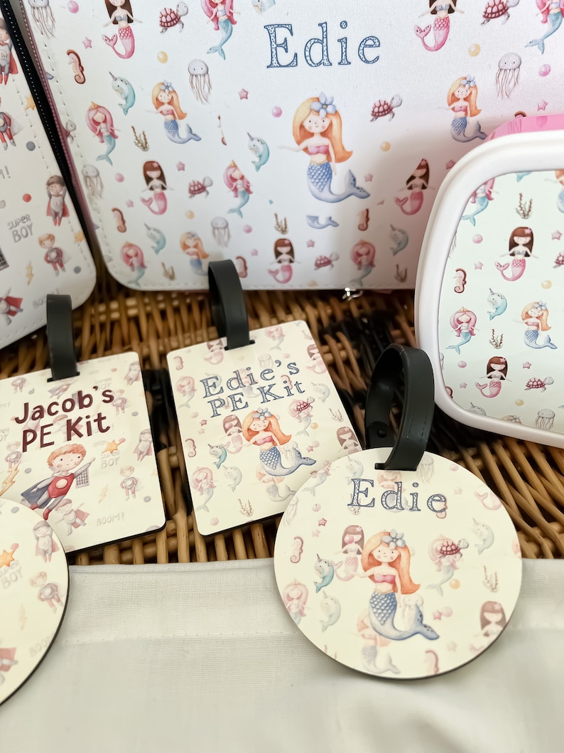 May include: Three personalized luggage tags with a mermaid theme. The tags are made of wood and have a white background with a colorful mermaid design. The text on the tags reads "Jacob's PE Kit", "Edie's PE Kit", and "Edie".