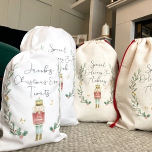 May include: Three white Christmas gift bags with red drawstrings. Each bag features a watercolor illustration of a nutcracker and a green wreath with gold stars. The bags are personalized with the names "Jacob's Christmas Eve Treats", "Special Delivery for Aubrey", and "Special Delivery for ...".