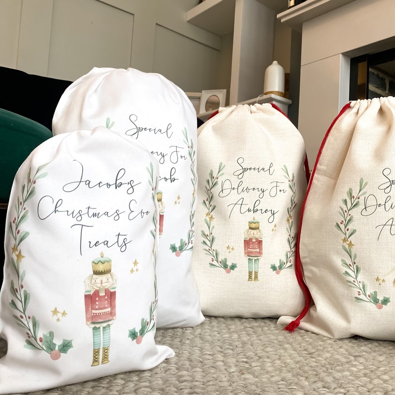 May include: Three white Christmas gift bags with red drawstrings. Each bag features a watercolor illustration of a nutcracker and a green wreath with gold stars. The bags are personalized with the names "Jacob's Christmas Eve Treats", "Special Delivery for Aubrey", and "Special Delivery for ...".