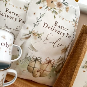 May include: Two white fabric Easter gift bags with bunny illustrations and floral designs. The bags are personalized with the text "Easter Delivery For Jacob" and "Easter Delivery For Eddie".