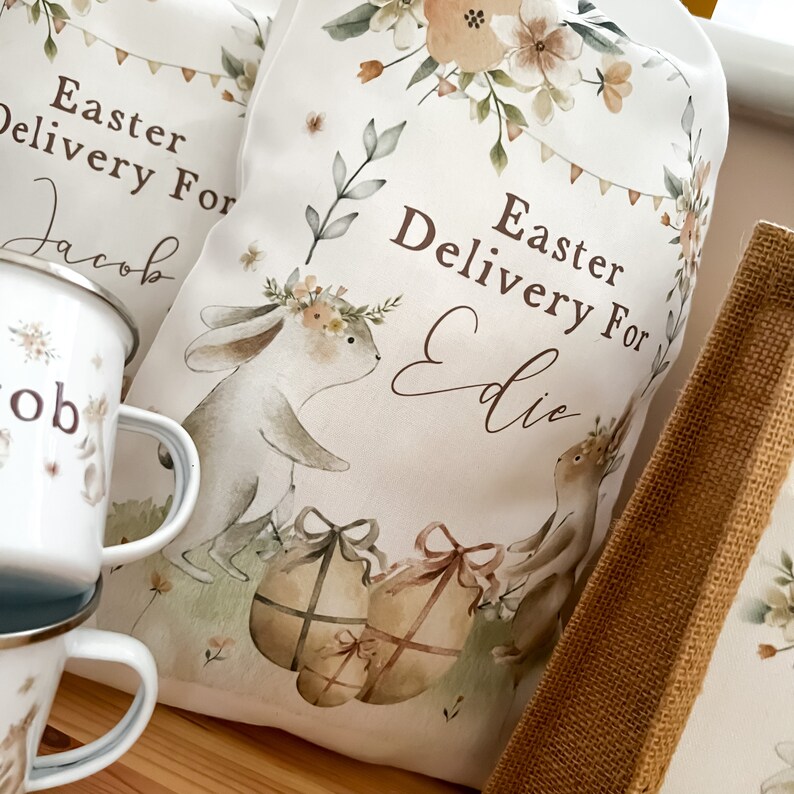 May include: Two white fabric Easter gift bags with bunny illustrations and floral designs. The bags are personalized with the text "Easter Delivery For Jacob" and "Easter Delivery For Eddie".