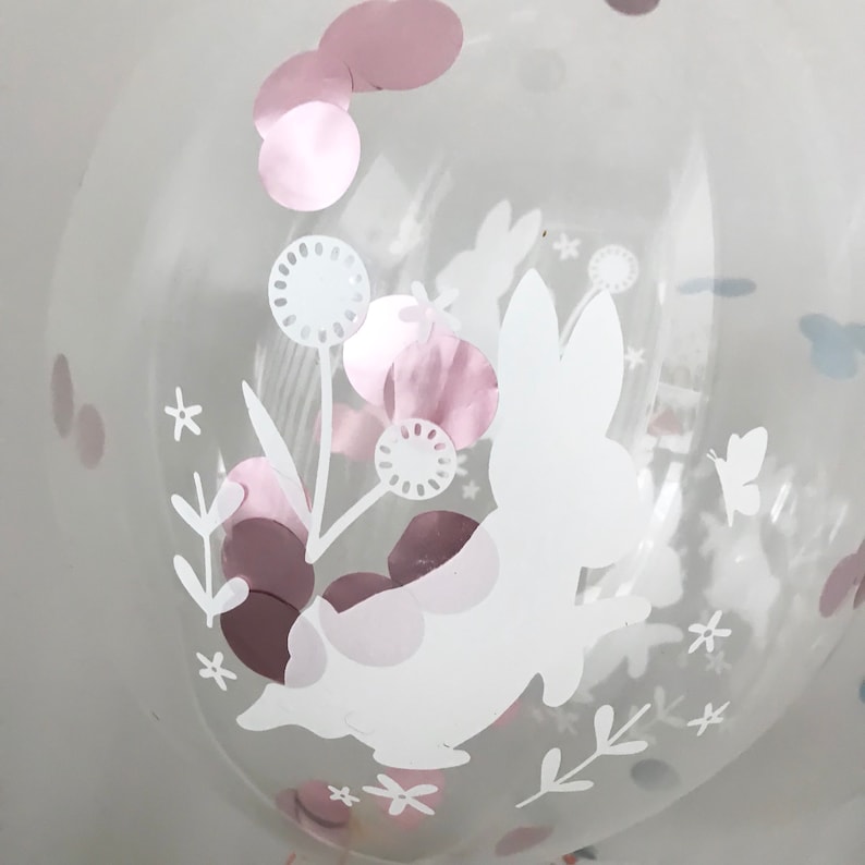 May include: A clear balloon with a white bunny rabbit design and pink and brown confetti. The bunny is holding a flower with a white stem and leaves.