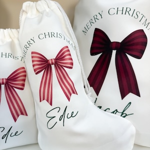 May include: White Christmas gift bags featuring a red and white striped bow design. The bags are printed with "Merry Christmas" in a circular pattern. One bag is stocking-shaped and has the name "Edie" printed on it.