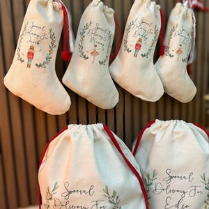 Pode incluir: Cinco sacos de presente de lona branca com cord&otilde;es de ajuste vermelhos. Cada saco tem um desenho de coroa verde com um personagem diferente no centro. Os sacos s&atilde;o etiquetados "Entrega especial para" seguido de um nome: Jacob, Mam&atilde;e, Papai, Edie e Jacob.