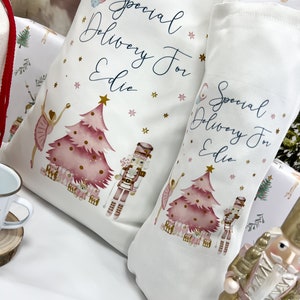 May include: Two white fabric stockings with pink and gold illustrations of a Christmas tree, a ballerina, and a nutcracker. The stockings have the text "Special Delivery For Edie" on them.