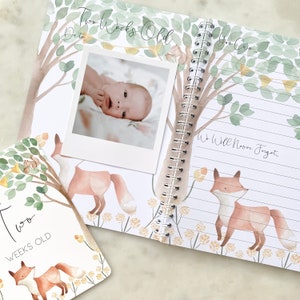 K&ouml;nnte beinhalten: Ein Baby-Erinnerungsbuch mit einer Aquarell-Illustration eines Fuchses und eines Baumes. Das Buch hat eine Spiralbindung und eine Seite mit einem Foto eines Babys. Der Text auf der Seite lautet "Zwei Wochen alt".
