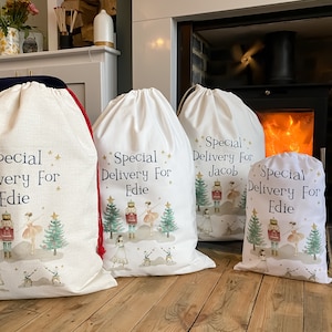 May include: Four white fabric gift bags with red drawstrings. Each bag has a different name printed on it: Edie, Edie, Jacob, and Edie. The bags feature a watercolour illustration of a nutcracker and a ballerina in front of a Christmas tree.