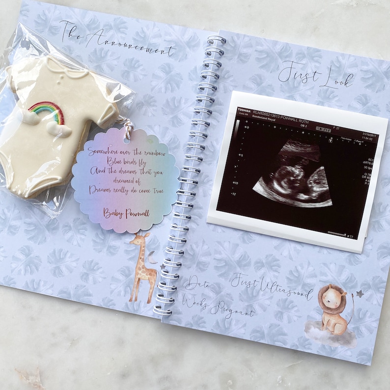 May include: A blue and white baby announcement scrapbook with a quote about dreams coming true, a cookie shaped like a onesie, and a first ultrasound image.