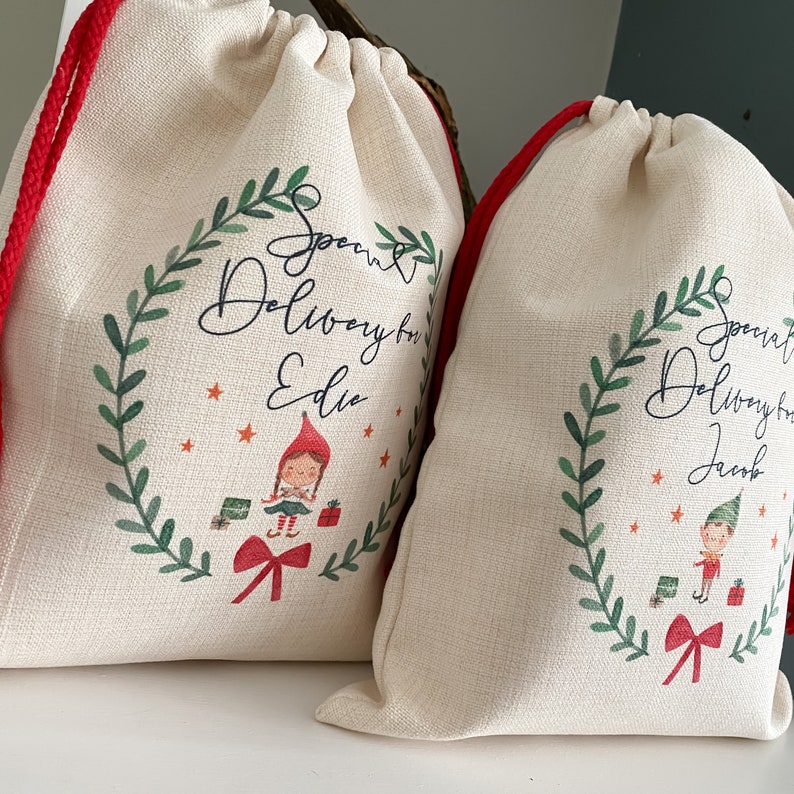 May include: Two white canvas gift bags with red drawstrings. The bags have a green wreath design with a red bow and a cartoon elf. The text on the bags reads "Special Delivery for [name]"