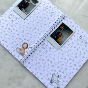 May include: A pregnancy journal with a white background and a pattern of stars and moons. The journal has a spiral binding and is open to pages 14, 15, 16, and 17. Each page has a space for a photo and a section for writing. The pages are decorated with illustrations of a lion, an elephant, and a baby.