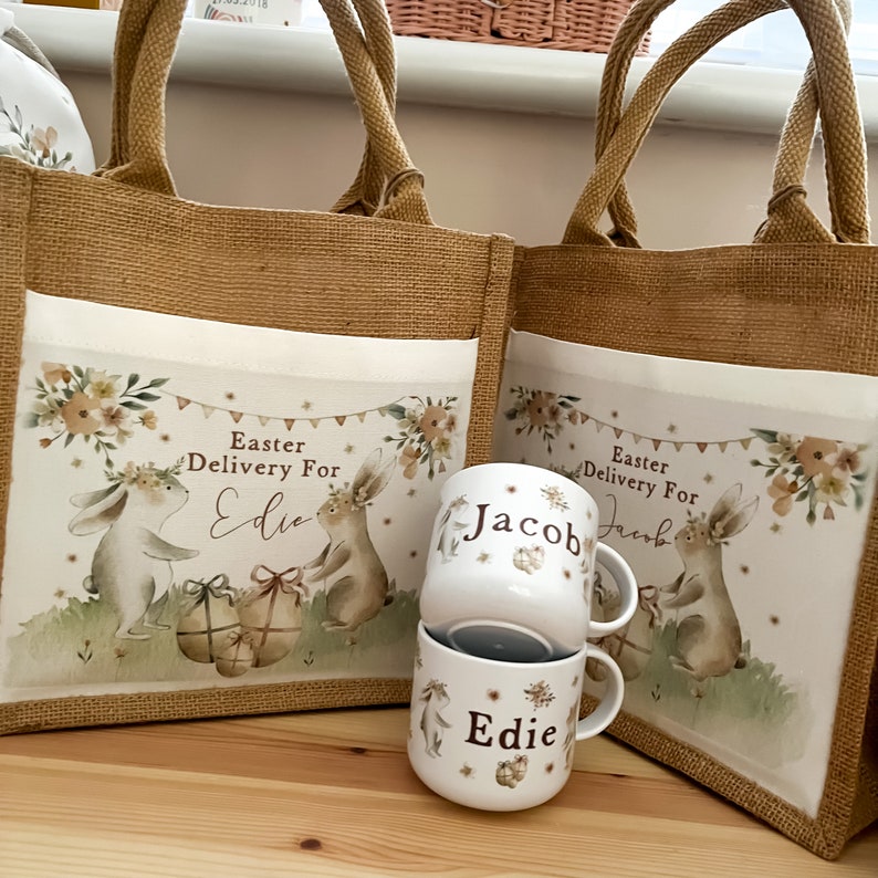 May include: Two burlap tote bags with personalized Easter bunny designs. The bags have the text "Easter Delivery For" and the names "Edie" and "Jacob". Two white ceramic mugs with the names "Edie" and "Jacob" are in front of the bags.