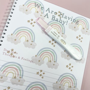 May include: A pregnancy journal with a rainbow and star pattern. The text "We Are Having A Baby!" is written on the cover. A positive pregnancy test is lying on the journal.