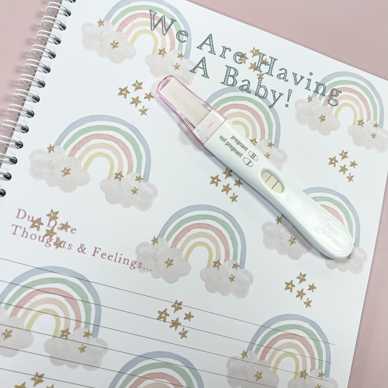 May include: A pregnancy journal with a rainbow and star pattern. The text "We Are Having A Baby!" is written on the cover. A positive pregnancy test is lying on the journal.