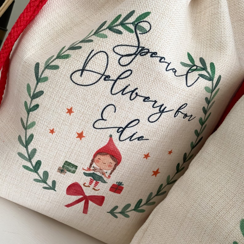 May include: A white canvas bag with a red drawstring. The bag has a green wreath design with the text "Special Delivery for Ellie" and a cartoon elf holding a gift.