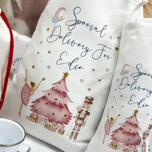 May include: Two white fabric gift bags with pink and gold illustrations of a ballerina, a Christmas tree, and a nutcracker. The bags have the text "Special Delivery For Edie" on them.