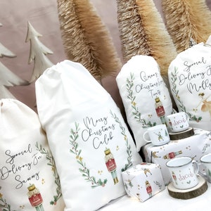 May include: Four white fabric gift bags with a Christmas theme. Each bag has a different design featuring a nutcracker, a ballerina, and a bottle of wine. The bags are decorated with green leaves, gold stars, and red berries. The bags are labeled with the names "Sammy", "Caleb", "Molly", and "Tints".