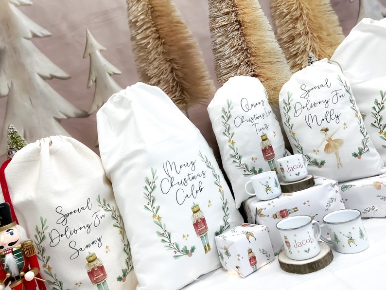 May include: Four white fabric gift bags with a Christmas theme. Each bag has a different design featuring a nutcracker, a ballerina, and a bottle of wine. The bags are decorated with green leaves, gold stars, and red berries. The bags are labeled with the names "Sammy", "Caleb", "Molly", and "Tints".