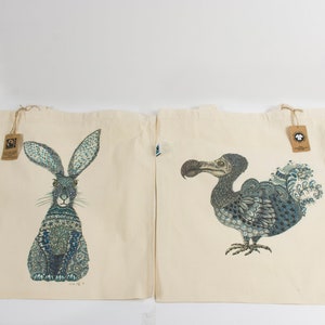 May include: Two beige tote bags with blue and black animal illustrations. One bag features a rabbit with blue and black patterned fur. The other bag features a dodo bird with blue and black patterned feathers.